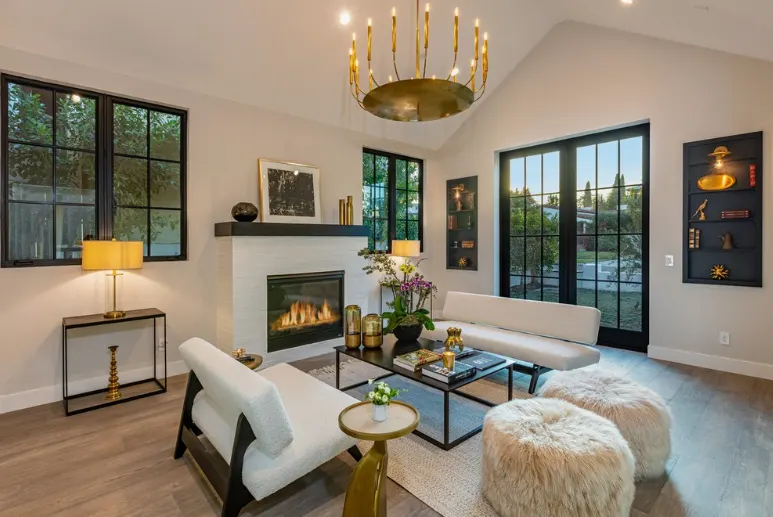 Bright remodeled living room with fireplace, high ceiling, chandelier, and large black-framed windows.